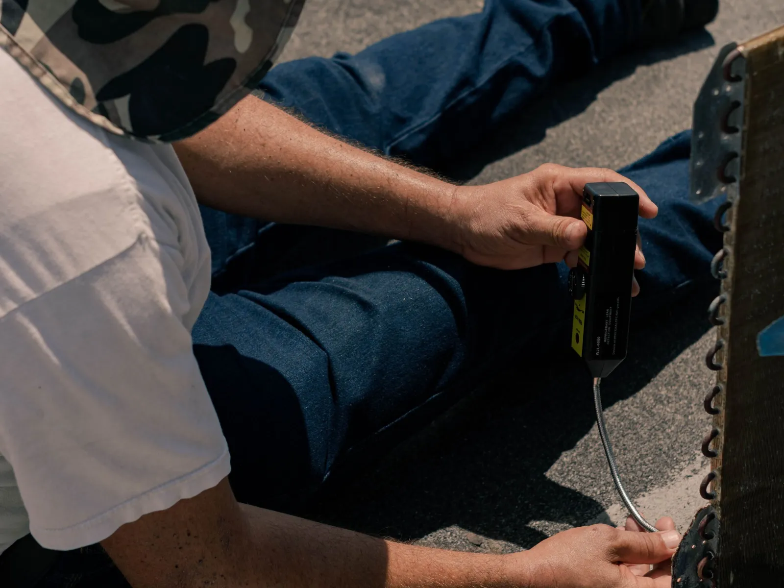 Seated technician checking an air-conditioner coil with a handheld refrigerant leak detector