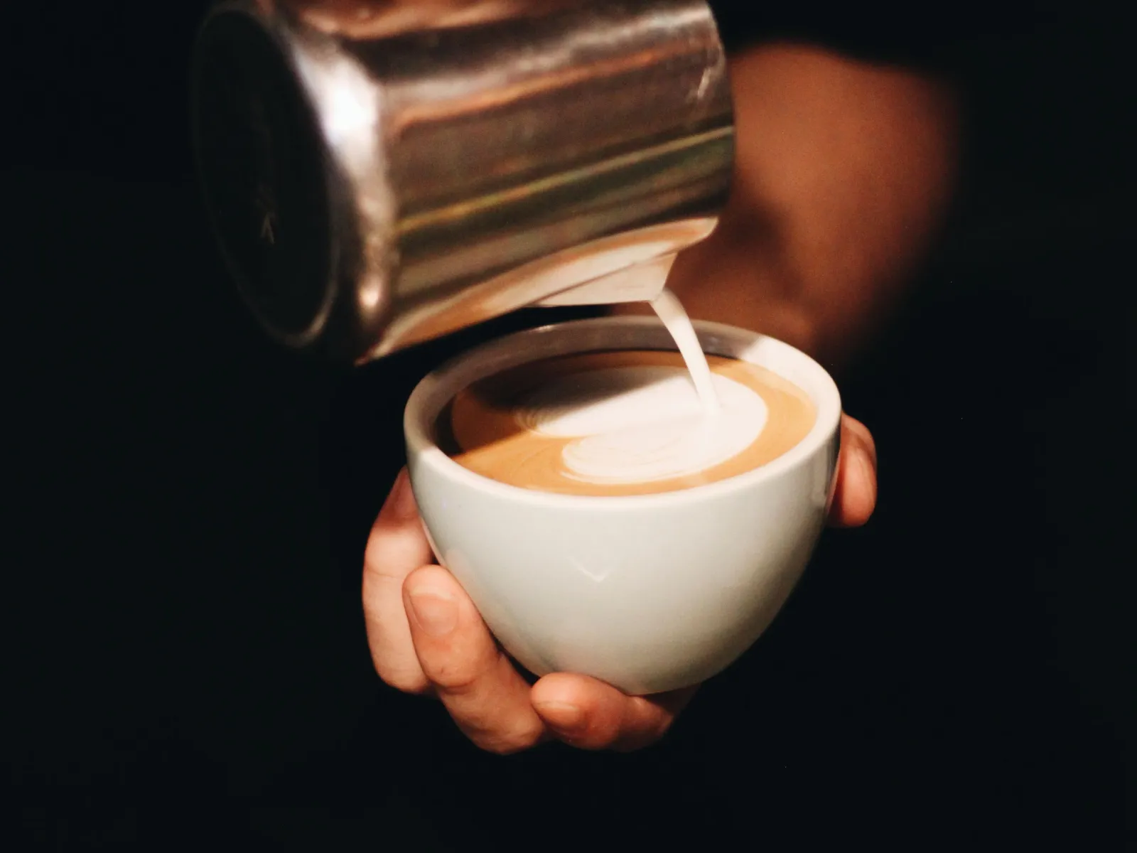 Hands pouring steamed milk from a steel pitcher into a coffee cup to form latte art