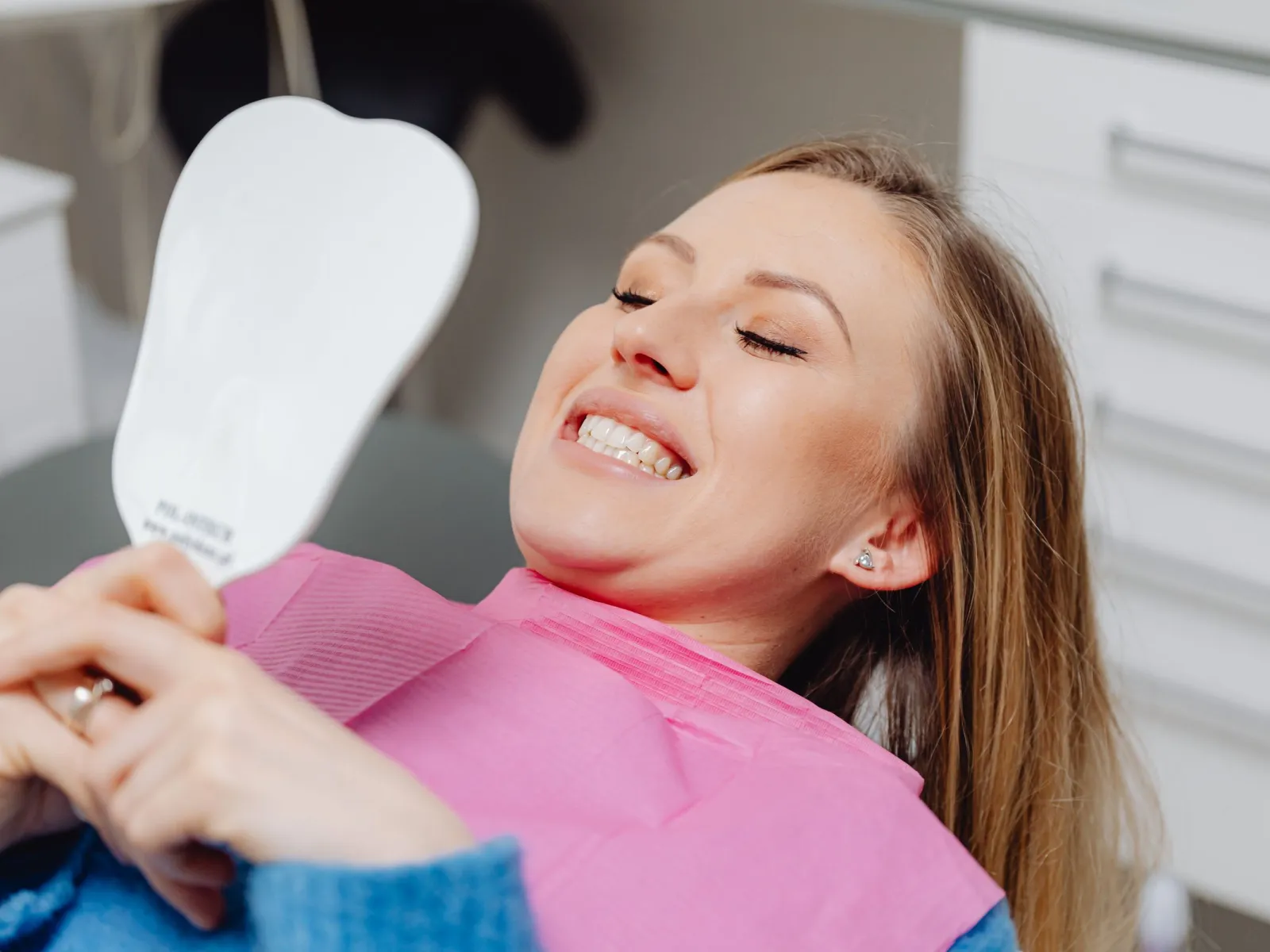 Smiling patient in a dental chair checking her teeth with a tooth-shaped mirror