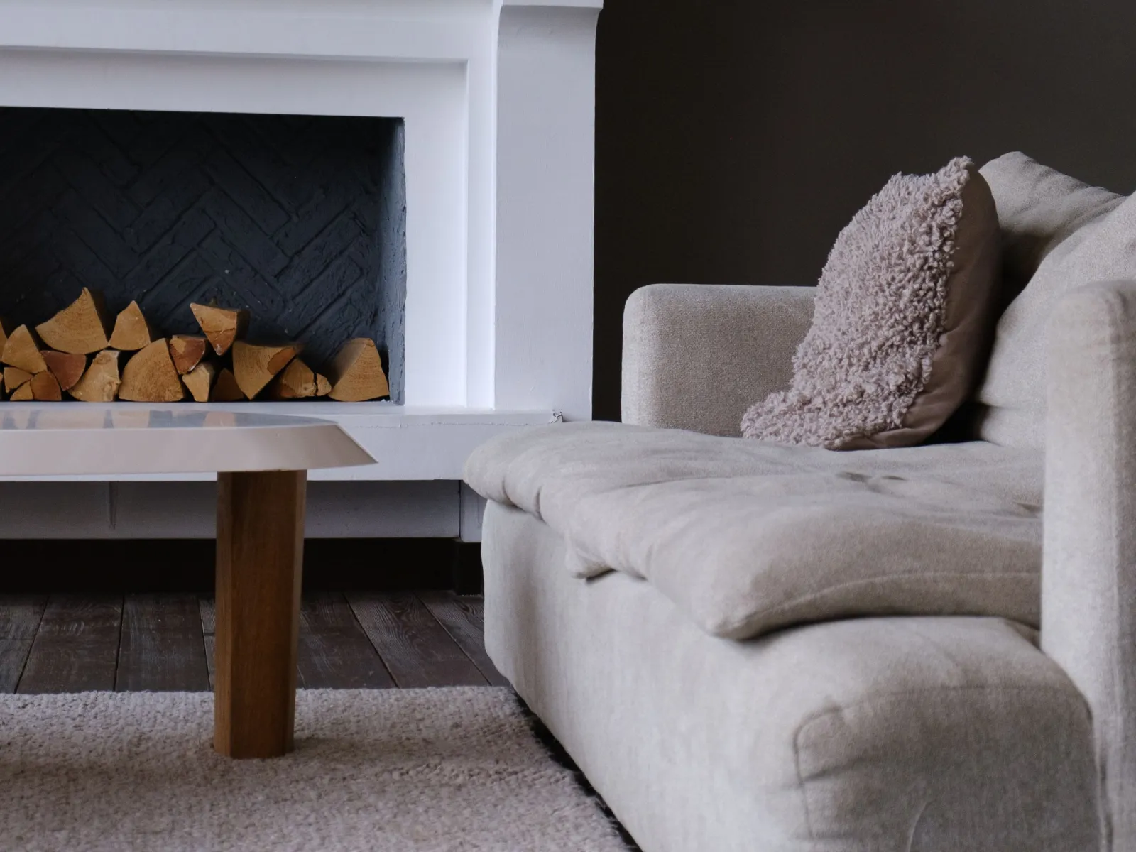 Beige fabric sofa beside a white fireplace mantel stacked with firewood against a dark brown wall