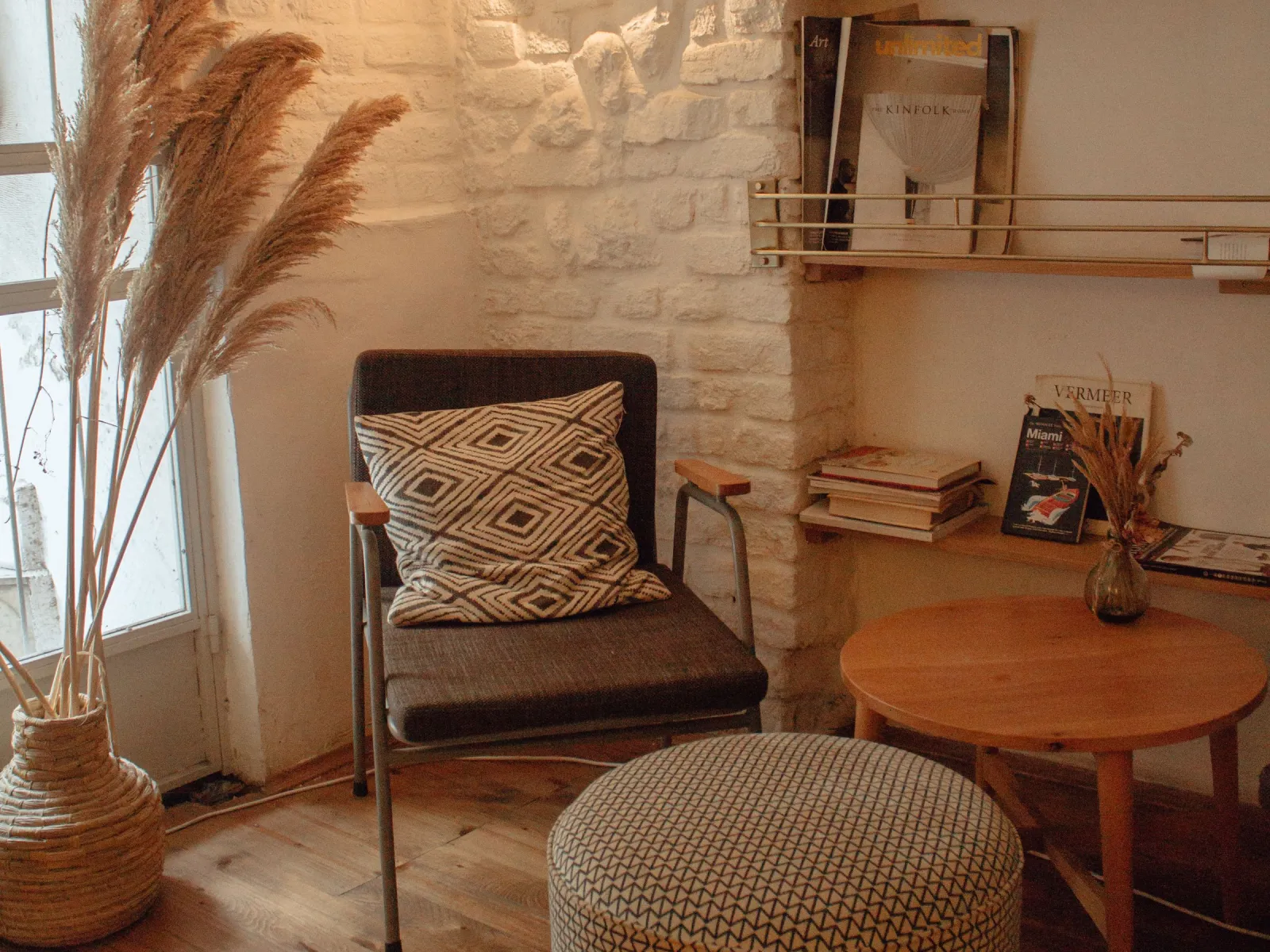 Cozy reading corner with dark armchair, patterned pouf, pampas grass and whitewashed brick wall