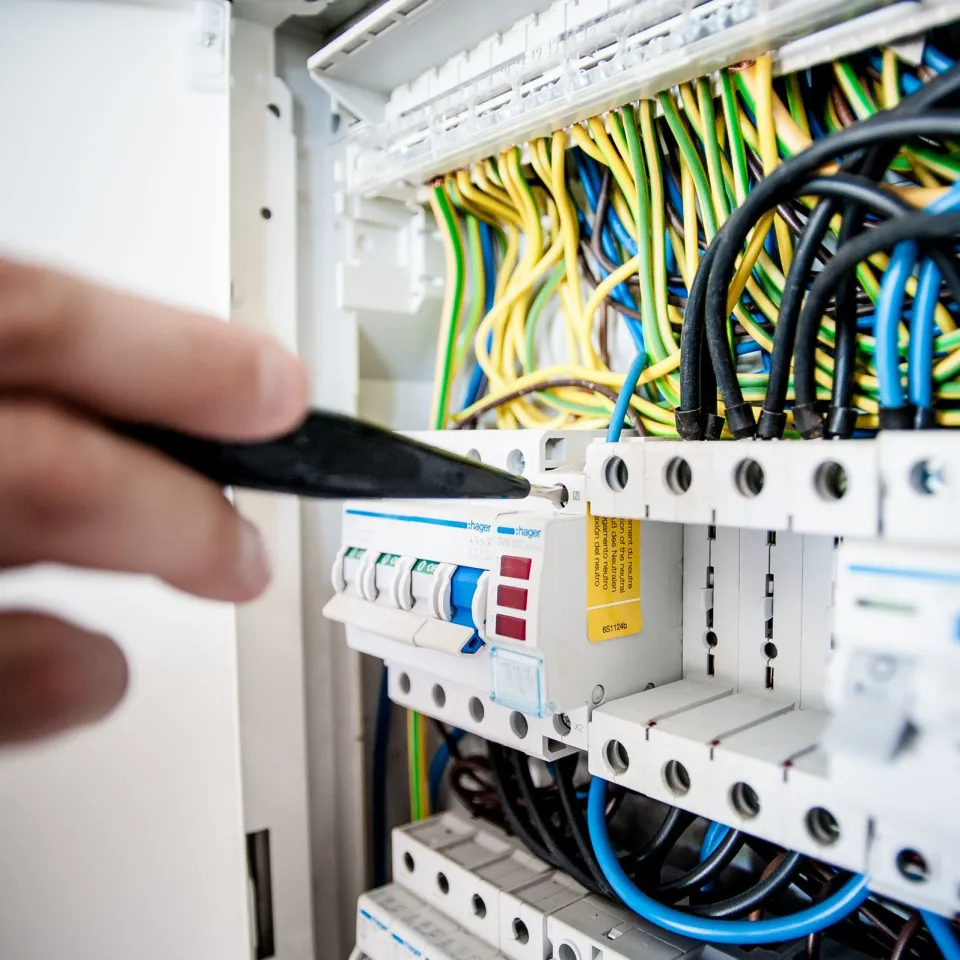 Hand guiding a screwdriver to a breaker terminal in a wiring panel full of color-coded cables
