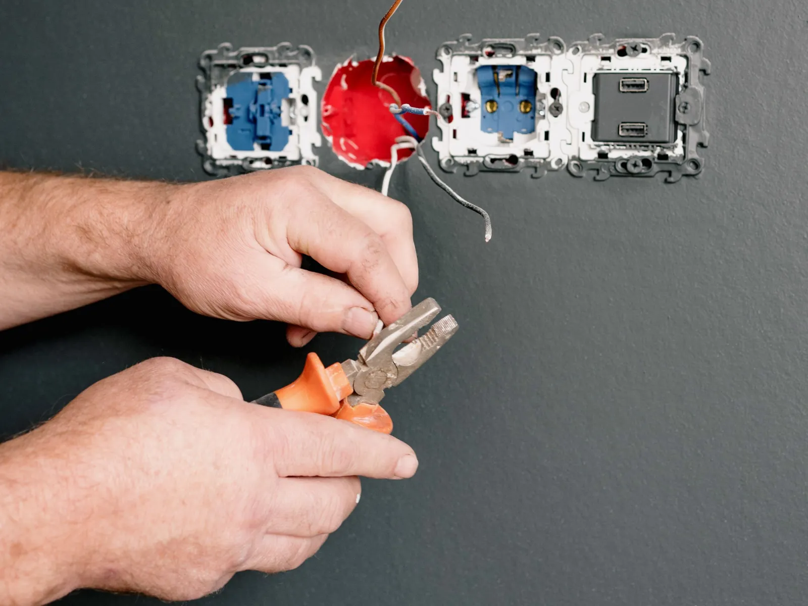 Electrician's hands using pliers on exposed wires below partly installed wall sockets