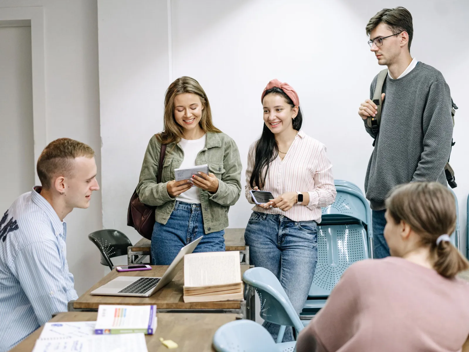 Five students chatting around a desk with a laptop and open book in a white classroom