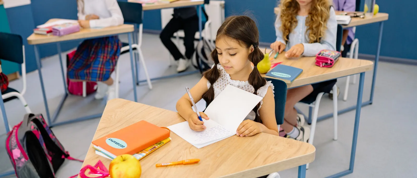 Primary students writing in notebooks at desks in a bright blue and white classroom